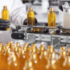 Plastic bottles with yellow liquid substance of shampoo, shower gel or soap standing in rows on assembly line at factory. Researcher in black gloves working at production line in modern laboratory